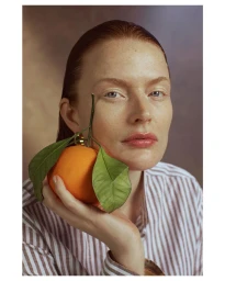 Portrait of a woman holding an orange with leaves by Marta Bevacqua, wearing a striped shirt against a warm textured background