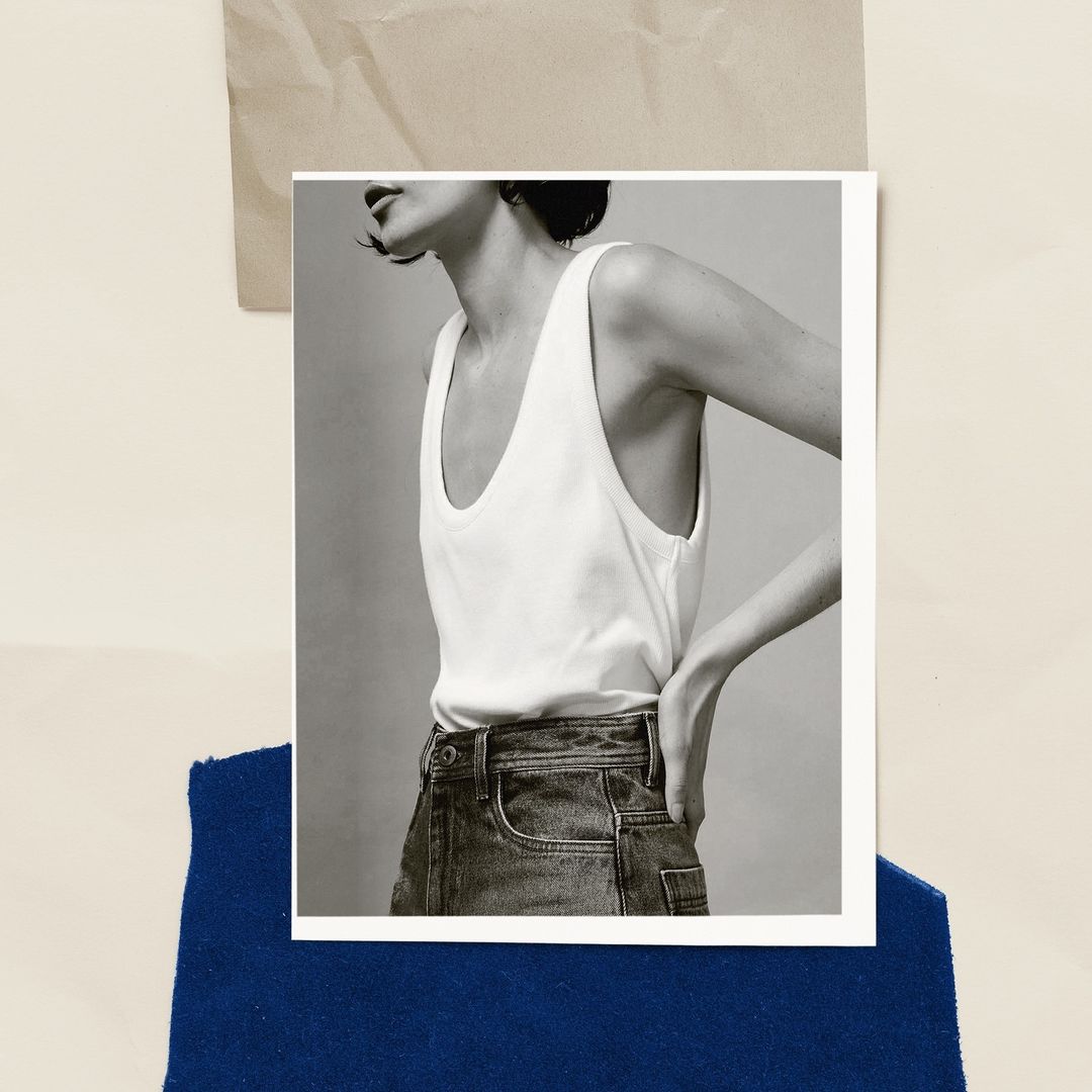 Monochrome fashion shot of a woman in a white tank top and denim jeans against a backdrop of blue fabric and neutral paper.