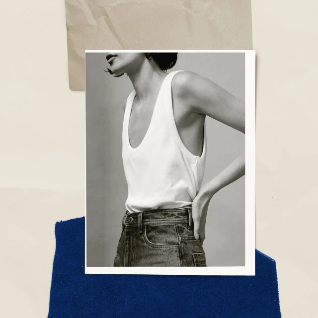 Monochrome fashion shot of a woman in a white tank top and denim jeans against a backdrop of blue fabric and neutral paper.