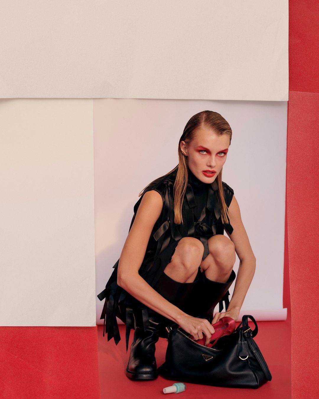 Fashion shot of a model in black clothing squatting with a Prada bag in a photo studio with red and white backdrops.
