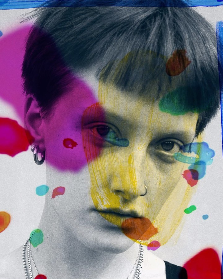 Close-up portrait of a person with short dark hair overlaid with vibrant paint splatters, silver hoop earring and chain necklace.