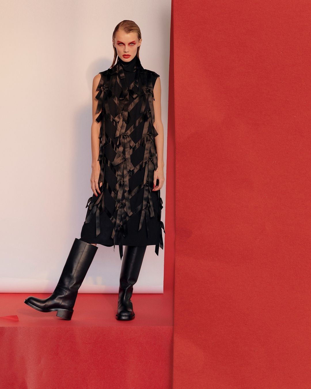 A fashion-forward adult female poses in a black dress with a geometric vest, knee-high boots, and dramatic red makeup in a studio setting.