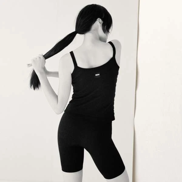 Monochrome portrait of a woman with dark hair in a ponytail, wearing black athletic top and shorts, back to viewer against a light wall.