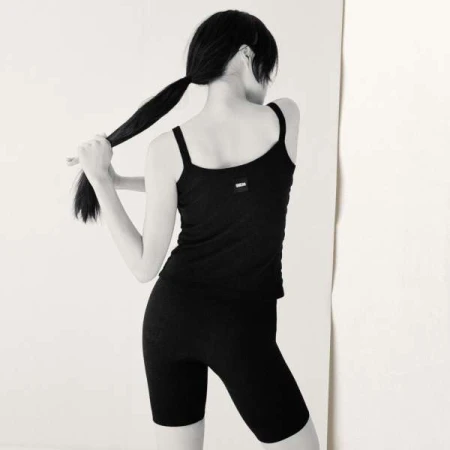 Monochrome portrait of a woman with dark hair in a ponytail, wearing black athletic top and shorts, back to viewer against a light wall.