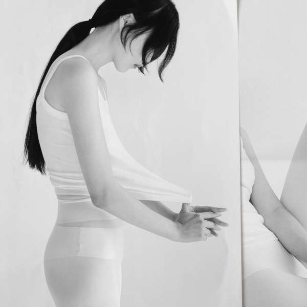 Monochrome portrait of a slim adult woman in a white tank top, side view, looking down, hands clasped, reflected on the right side.