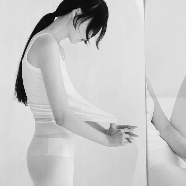 Monochrome portrait of a slim adult woman in a white tank top, side view, looking down, hands clasped, reflected on the right side.