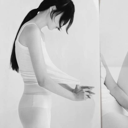 Monochrome portrait of a slim adult woman in a white tank top, side view, looking down, hands clasped, reflected on the right side.