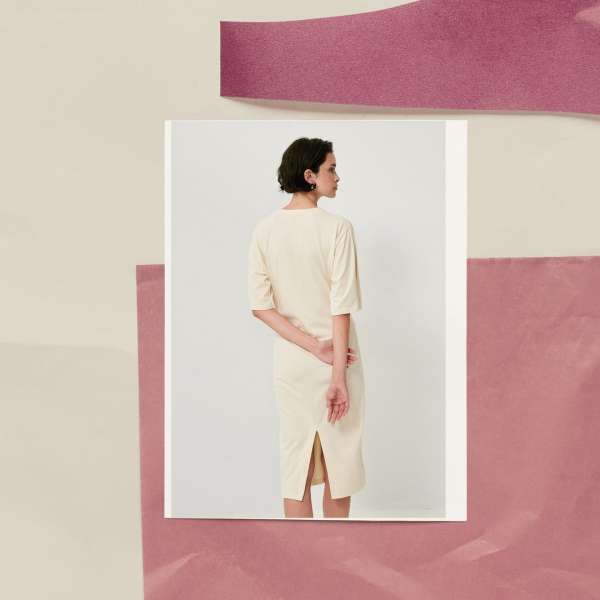 Woman in cream dress with short dark hair and hands clasped behind back, surrounded by paper collages in pink and cream tones.