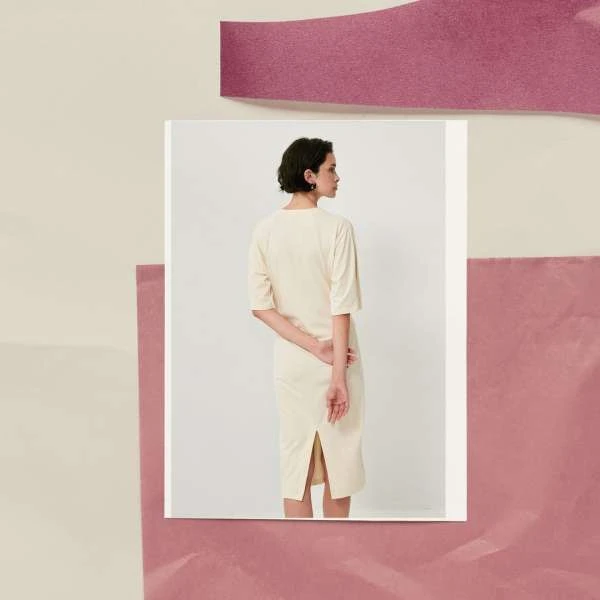Woman in cream dress with short dark hair and hands clasped behind back, surrounded by paper collages in pink and cream tones.