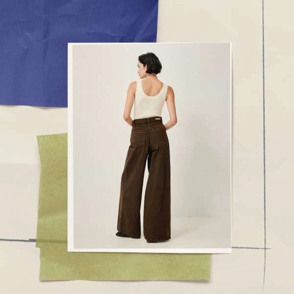 A woman models wide-legged, dark-brown jeans and a cream-colored tank top against a layered paper art background in a studio setting.
