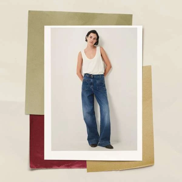Full shot of an adult woman wearing a tank top and wide-leg jeans, leaning against a white wall, with layered colored paper behind her.