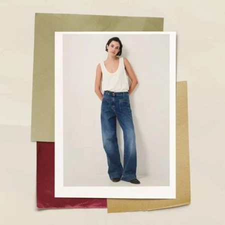 Full shot of an adult woman wearing a tank top and wide-leg jeans, leaning against a white wall, with layered colored paper behind her.