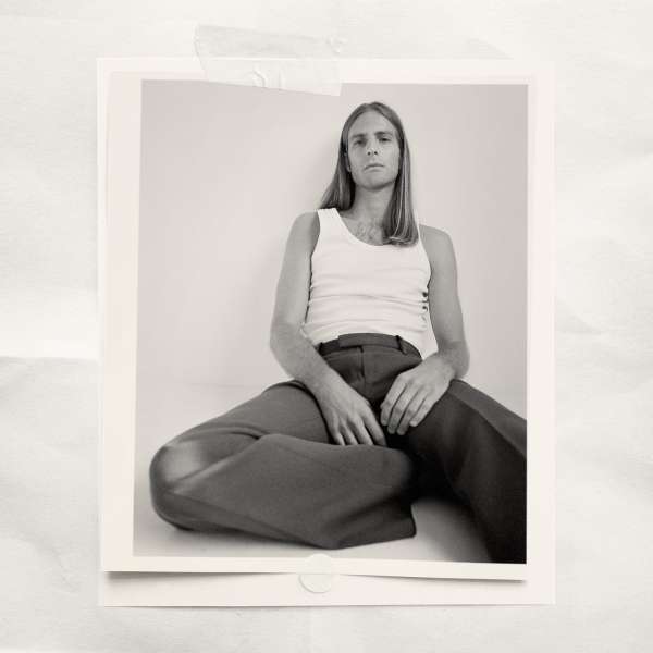 Portrait of an adult male in a white tank top and brown pants seated on a white floor in a photo studio with neutral lighting.