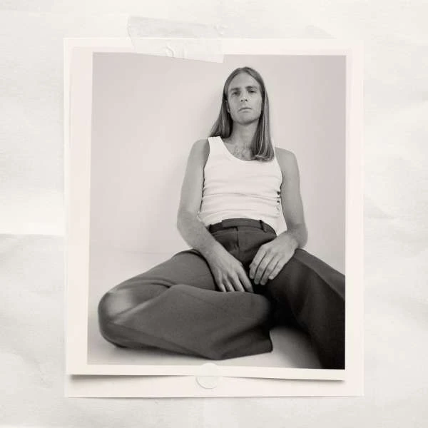 Portrait of an adult male in a white tank top and brown pants seated on a white floor in a photo studio with neutral lighting.