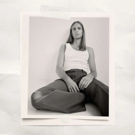 Portrait of an adult male in a white tank top and brown pants seated on a white floor in a photo studio with neutral lighting.