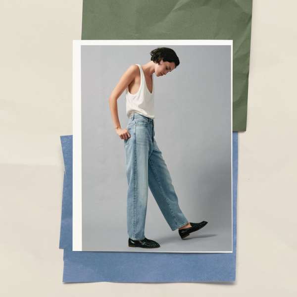 A full-shot fashion photograph features a woman in a white tank top, jeans, and loafers on a white card with a paper background.