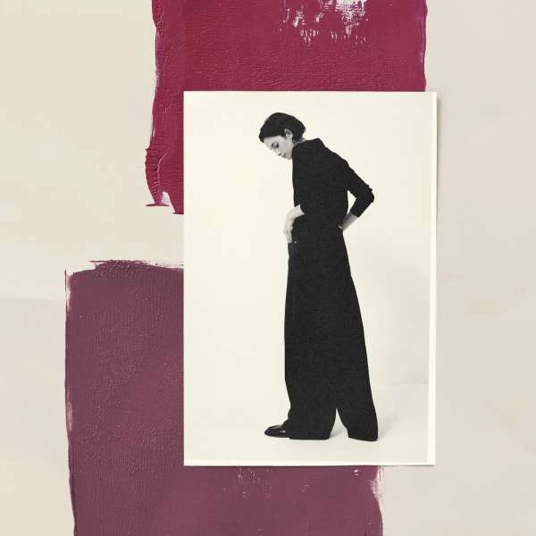 A woman in black wide-leg pants and top stands on a white surface against an abstract painted backdrop of cream and burgundy hues.