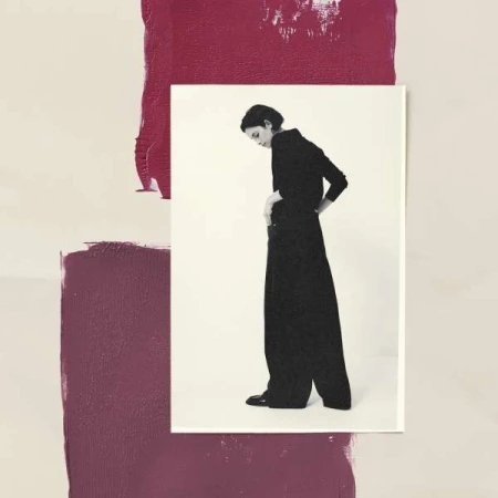 A woman in black wide-leg pants and top stands on a white surface against an abstract painted backdrop of cream and burgundy hues.