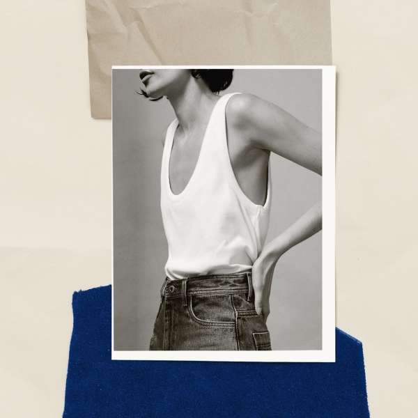 Black-and-white fashion shot of a young adult woman wearing a white tank top and blue jeans against a neutral studio backdrop.