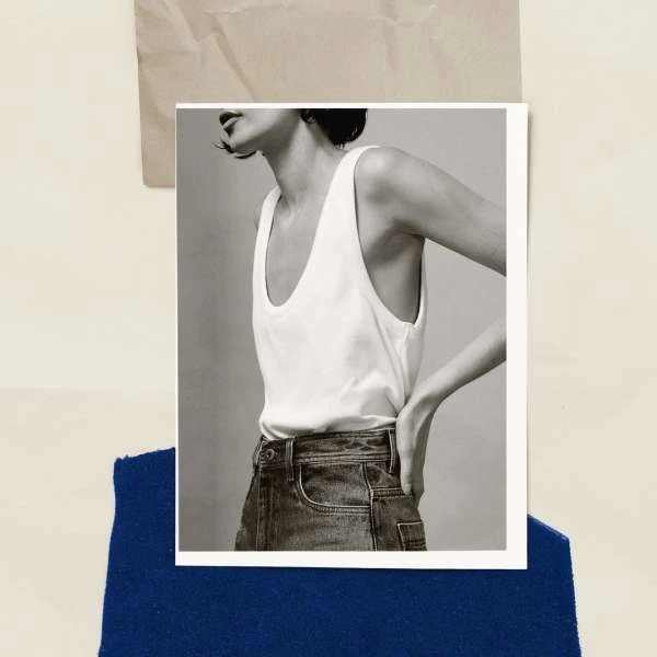 Black-and-white fashion shot of a young adult woman wearing a white tank top and blue jeans against a neutral studio backdrop.
