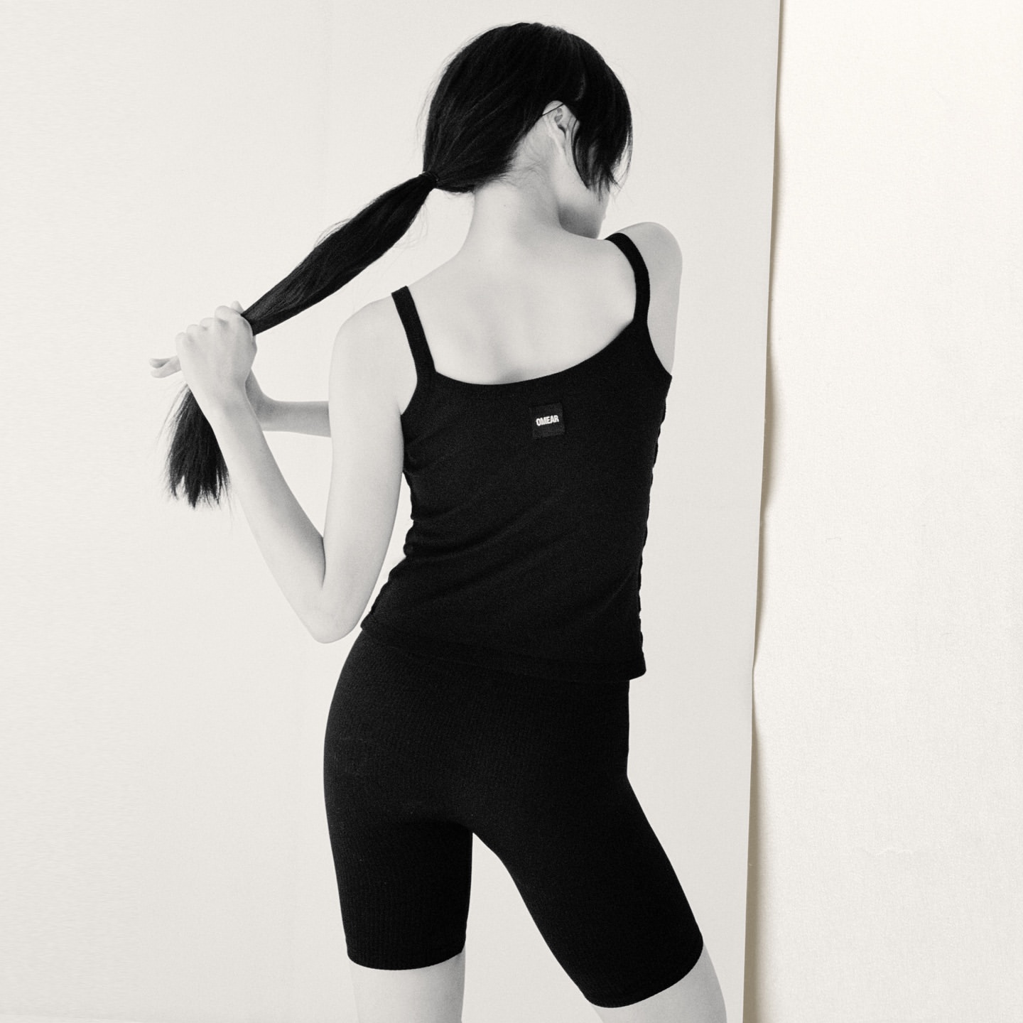 Back view of a young adult woman with dark hair in a ponytail wearing a black ribbed tank top and bike shorts against a white backdrop.