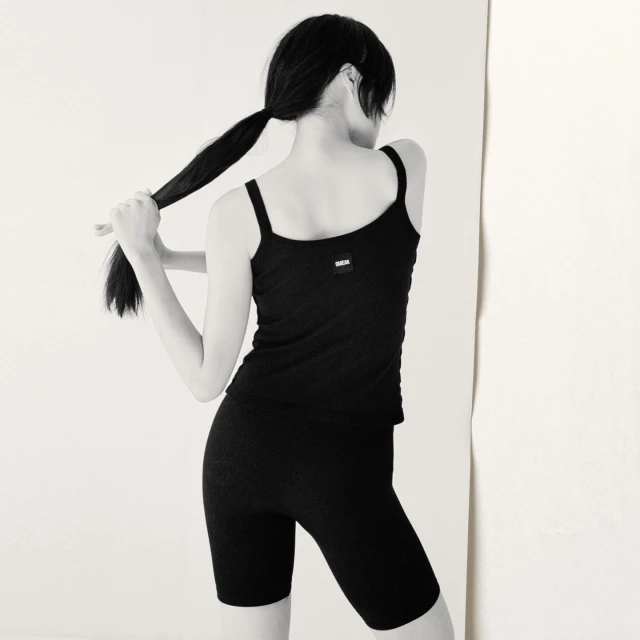 Back view of a young adult woman with dark hair in a ponytail wearing a black ribbed tank top and bike shorts against a white backdrop.