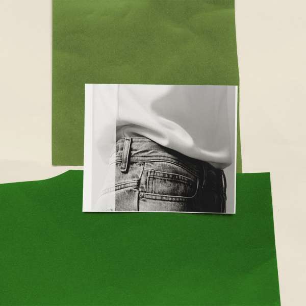 Cropped black and white photo of person wearing blue jeans, against green and beige paper sheets in a studio lighting setup.