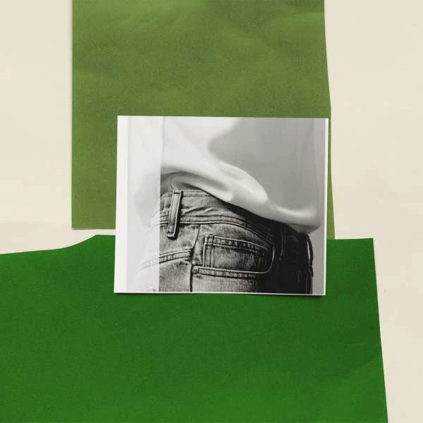 Cropped black and white photo of person wearing blue jeans, against green and beige paper sheets in a studio lighting setup.