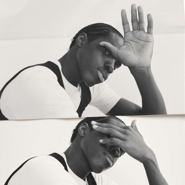Monochrome portrait of an adult man with braided hair, hands raised to his face, wearing a white t-shirt with black shoulder straps.