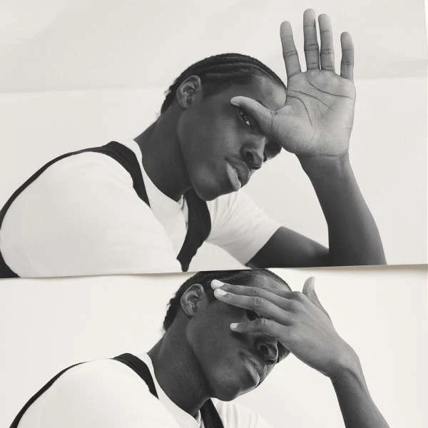 Monochrome portrait of an adult man with braided hair, hands raised to his face, wearing a white t-shirt with black shoulder straps.