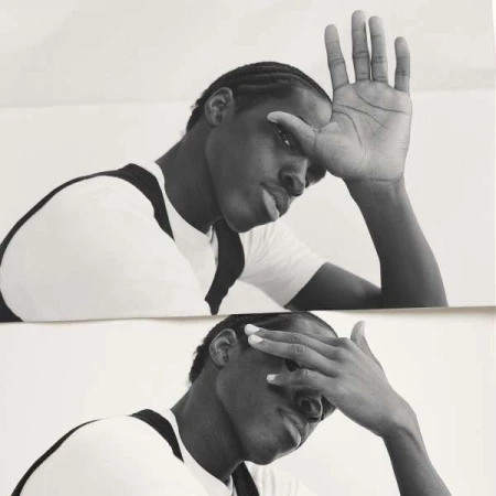 Monochrome portrait of an adult man with braided hair, hands raised to his face, wearing a white t-shirt with black shoulder straps.