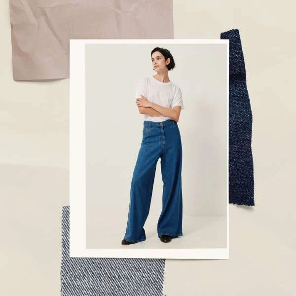 Woman in white shirt and wide-leg jeans posing indoors against a beige and denim textile backdrop for a fashion editorial.