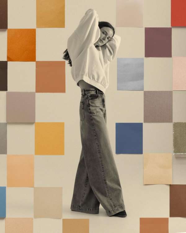A young woman stands in a photo studio against a colorful, grid-patterned backdrop, wearing a white sweatshirt and grey denim jeans.