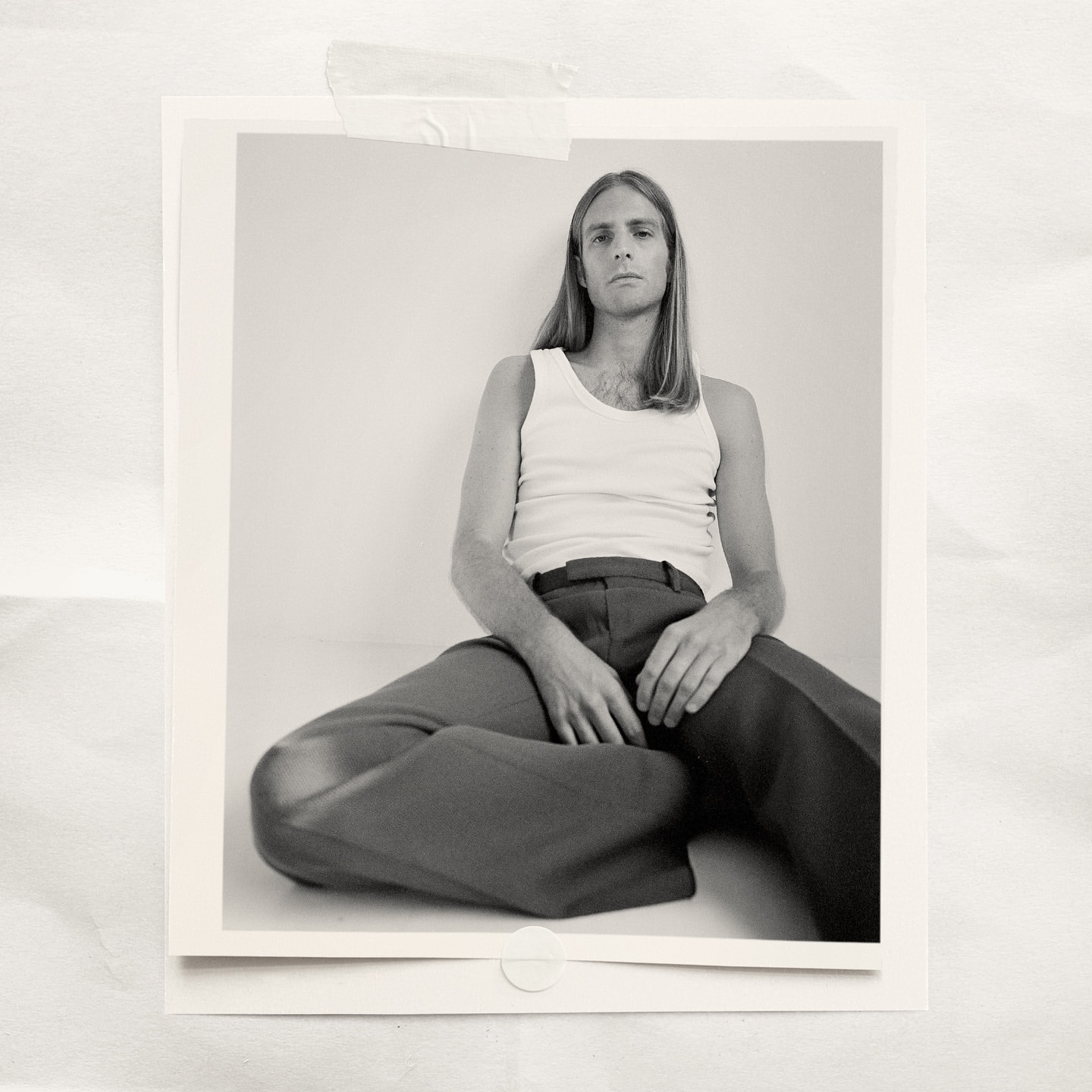 Portrait of a young adult male with long blonde hair, wearing a white tank top and dark pants, sitting indoors in front of a white wall.