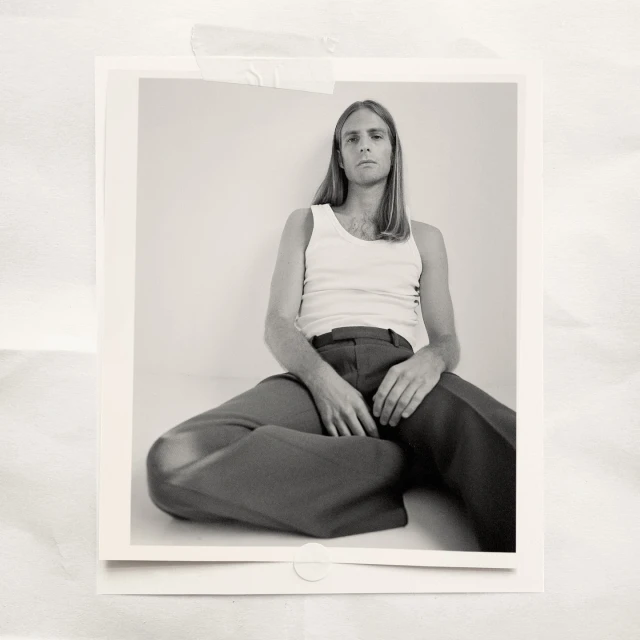 Portrait of a young adult male with long blonde hair, wearing a white tank top and dark pants, sitting indoors in front of a white wall.