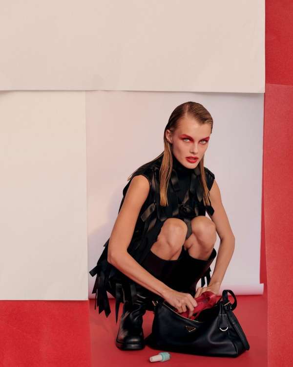 A woman in a black vest crouching with a black Prada handbag, red interior, white and red background, and a small tube on the floor.