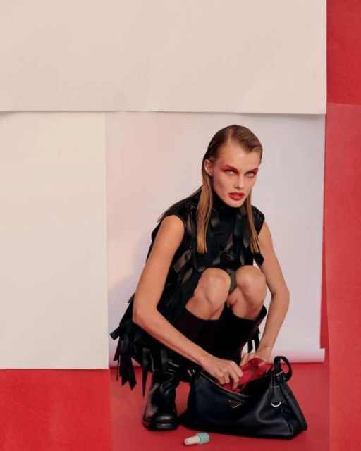 A woman in a black vest crouching with a black Prada handbag, red interior, white and red background, and a small tube on the floor.