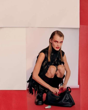 A woman in a black vest crouching with a black Prada handbag, red interior, white and red background, and a small tube on the floor.
