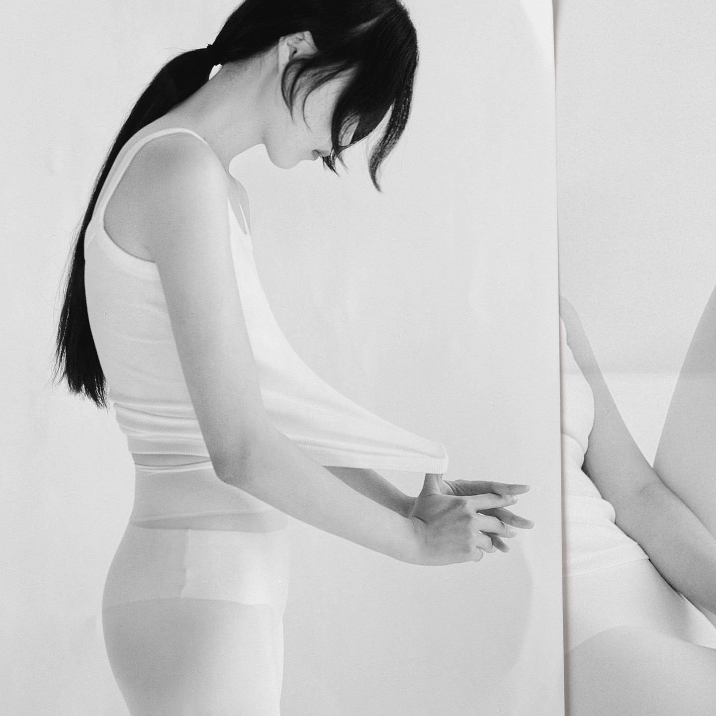 Black and white portrait of a slender adult female in a tank top and stockings, standing in a minimalist photo studio with a mirror.