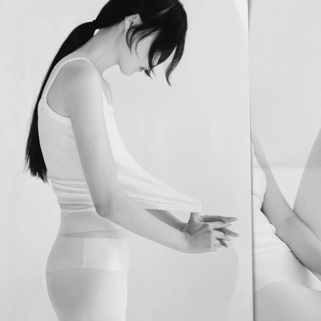 Black and white portrait of a slender adult female in a tank top and stockings, standing in a minimalist photo studio with a mirror.