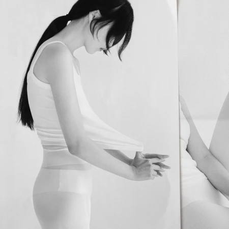 Black and white portrait of a slender adult female in a tank top and stockings, standing in a minimalist photo studio with a mirror.