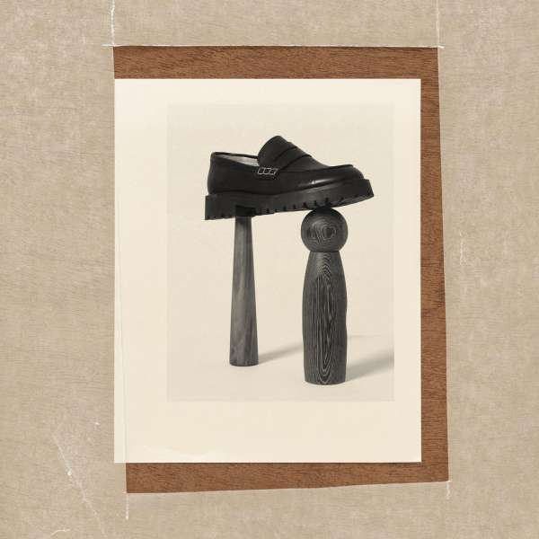 Still life captures a black leather loafer balanced atop dark wooden objects against a cream backdrop, framed by a brown border.