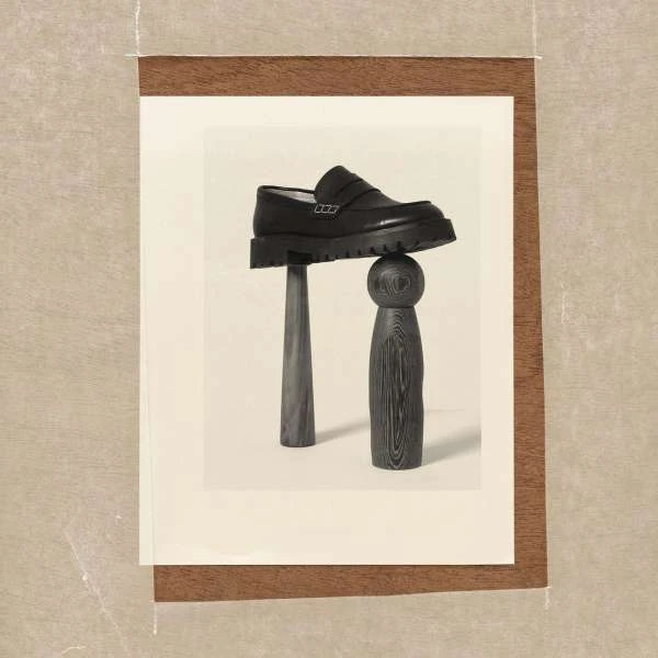 Still life captures a black leather loafer balanced atop dark wooden objects against a cream backdrop, framed by a brown border.