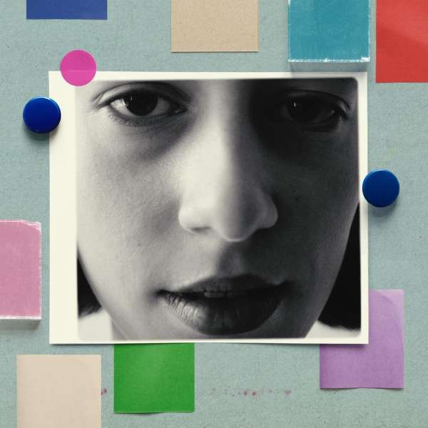 Close-up portrait of a woman's face in black and white, pinned to a light blue surface with colorful paper squares and magnets.
