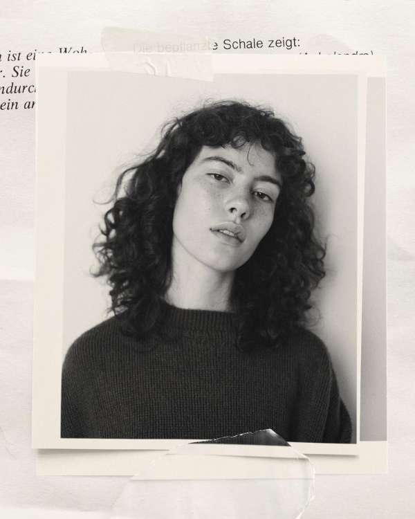 Monochrome portrait of a young adult woman with curly hair, wearing a dark sweater, set against a neutral background with paper textures.