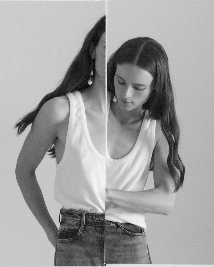 Black and white portrait of a woman in a white tank top and blue jeans, looking down with long hair and earrings, against a plain background.