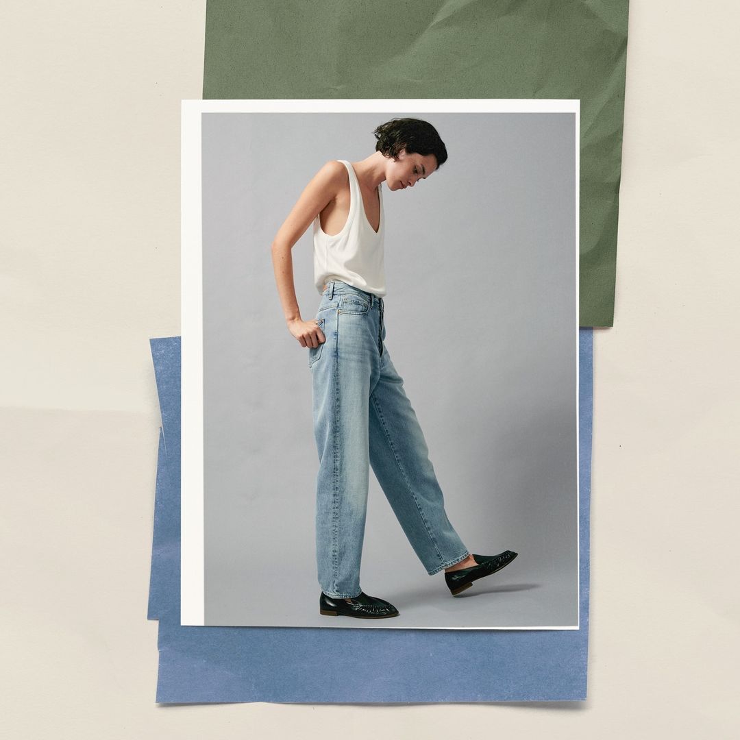 A slender woman in jeans and a tank top strikes a pose against a minimalist backdrop with layered paper accents and sleek black shoes.