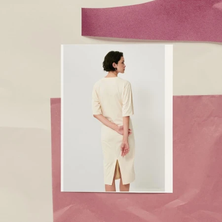 Woman in cream dress standing against a white square. The background is pink and cream. Fashion editorial with unique composition.
