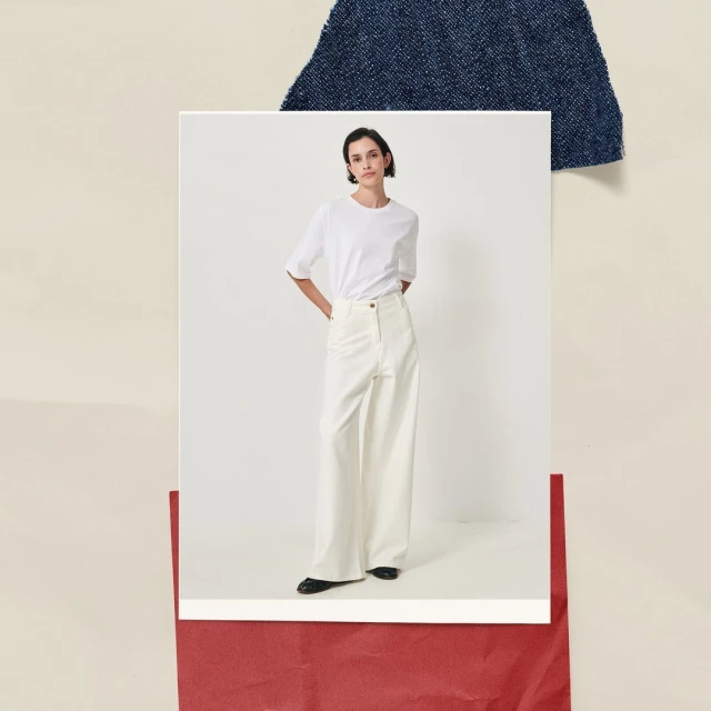 Studio shot of a woman in a white t-shirt and off-white wide-leg pants, standing against neutral backdrop with denim and red paper.