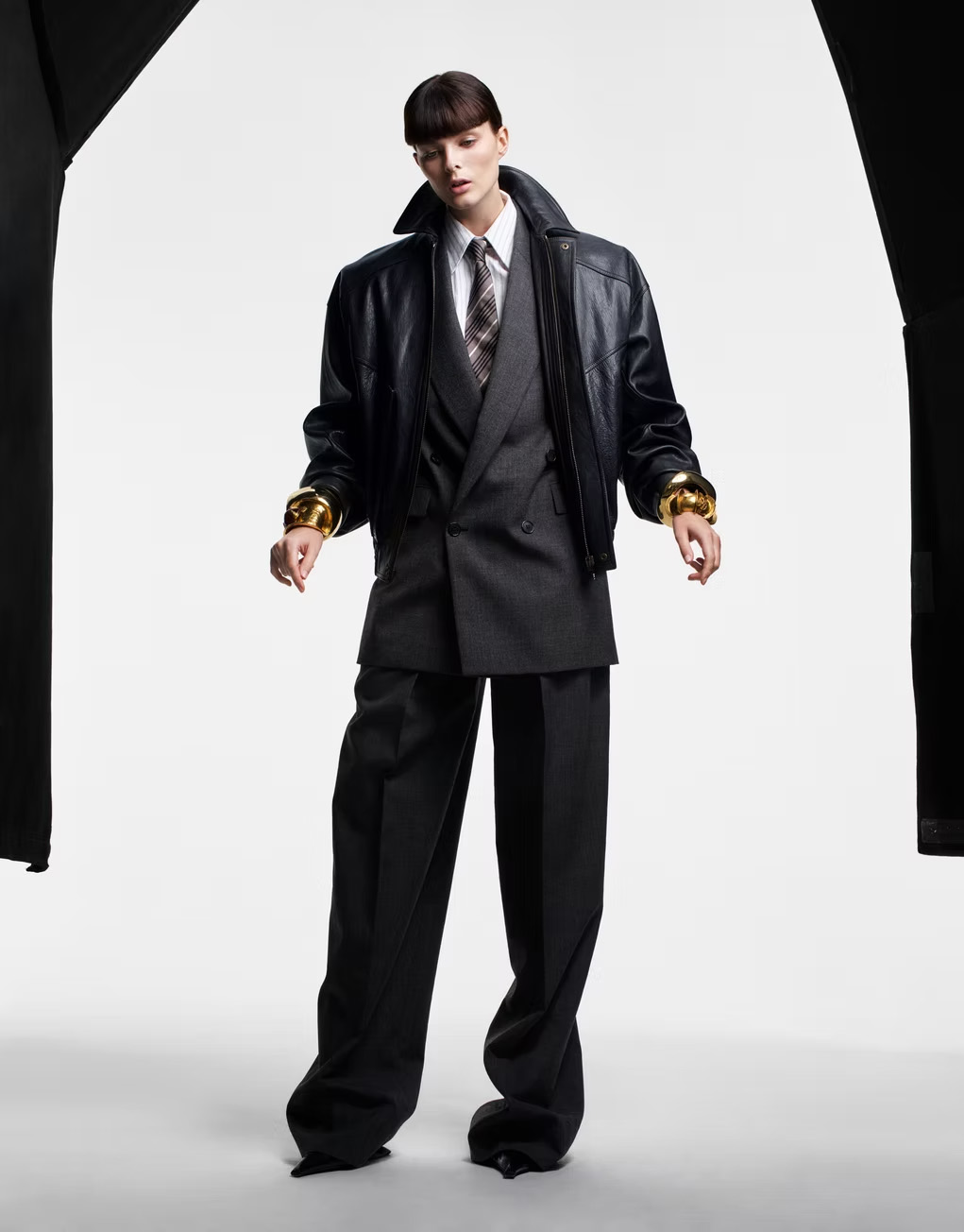 Full-length portrait of a woman in a grey suit, white shirt, tie, and black leather jacket, styled with gold bracelets against a white backdrop.